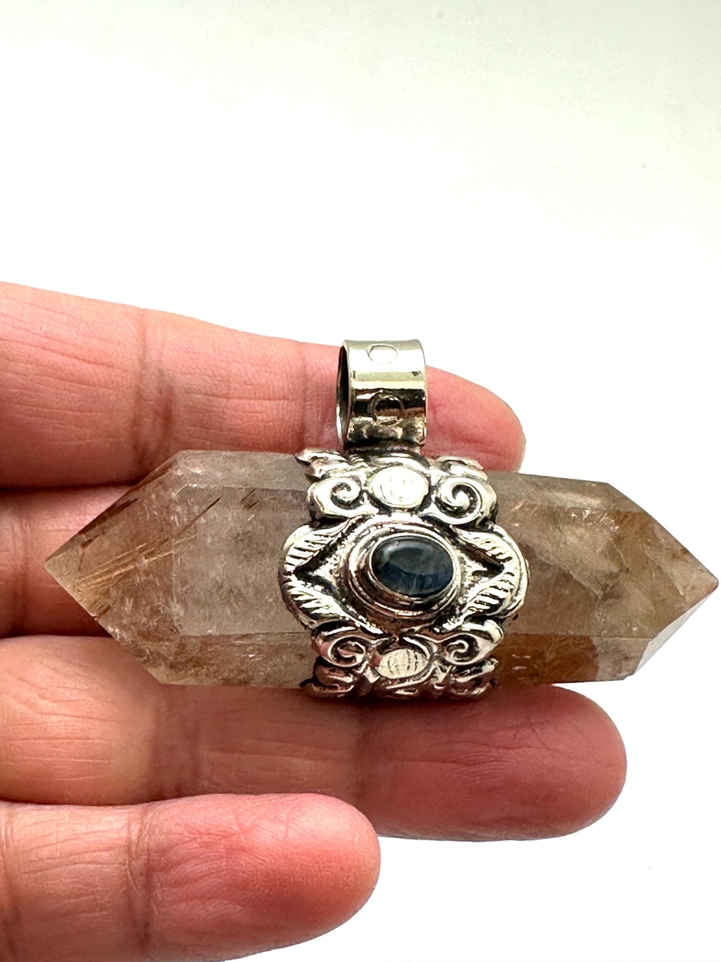 May include: A double-terminated quartz crystal pendant with a silver-colored metal setting. The crystal is a translucent brown color with internal inclusions. The setting features an ornate design with a small, round, dark blue stone in the center.