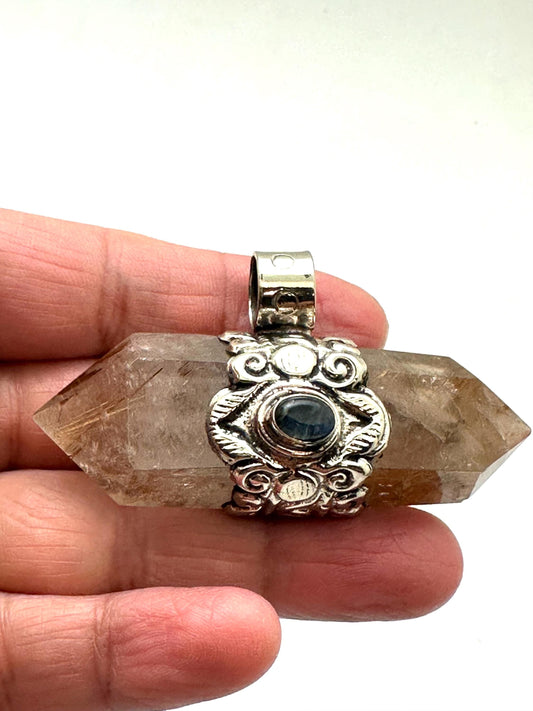 May include: A double-terminated quartz crystal pendant with a silver-colored metal setting. The crystal is a translucent brown color with internal inclusions. The setting features an ornate design with a small, round, dark blue stone in the center.